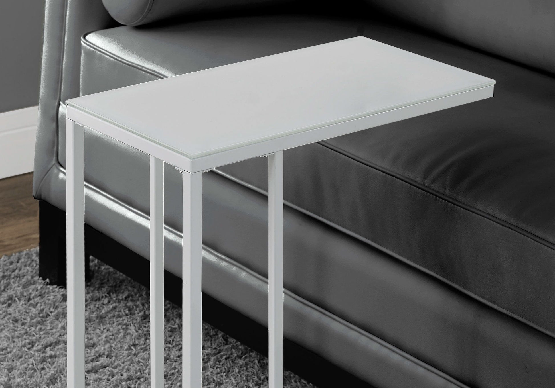 A C-shaped Accent Table with a tempered glass top and stylish contemporary design stands beside a dark gray leather sofa on a gray carpet, its minimalist metal frame positioned near the sofa armrest.