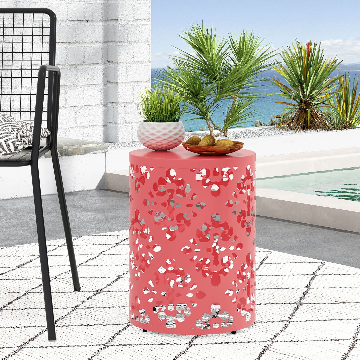The Outdoor Metal Modern Design End Table, coral pink and cylindrical with a cut-out leaf pattern, stands by a pool topped with decor. A modern chair, white patterned rug, lush greenery, and the ocean create a vibrant backdrop.