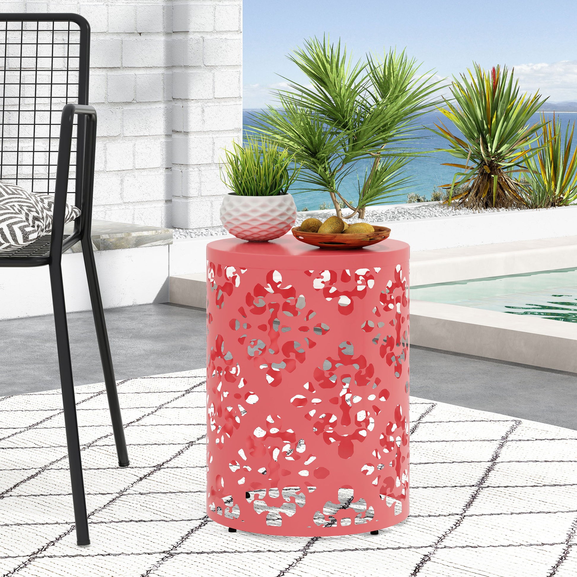 The Outdoor Metal Modern Design End Table, coral pink and cylindrical with a cut-out leaf pattern, stands by a pool topped with decor. A modern chair, white patterned rug, lush greenery, and the ocean create a vibrant backdrop.