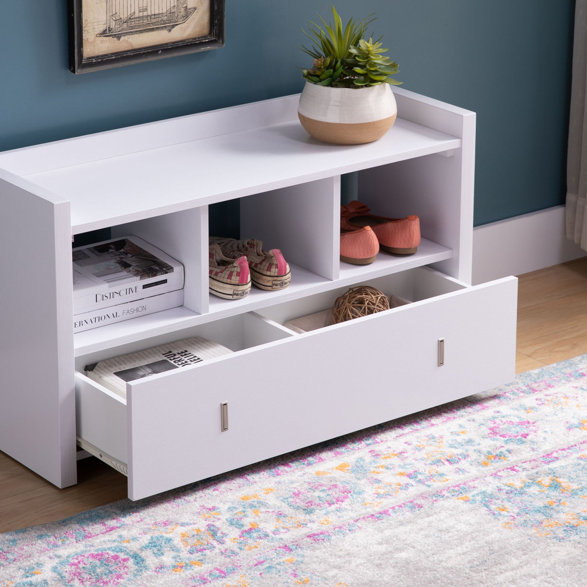 The Shoe Entry Bench With Three Shelves and One Drawer With Divided Compartment, in white, organizes shoes, books, a basket, and a plant. It stands on a wood floor beside a colorful rug and blue wall.