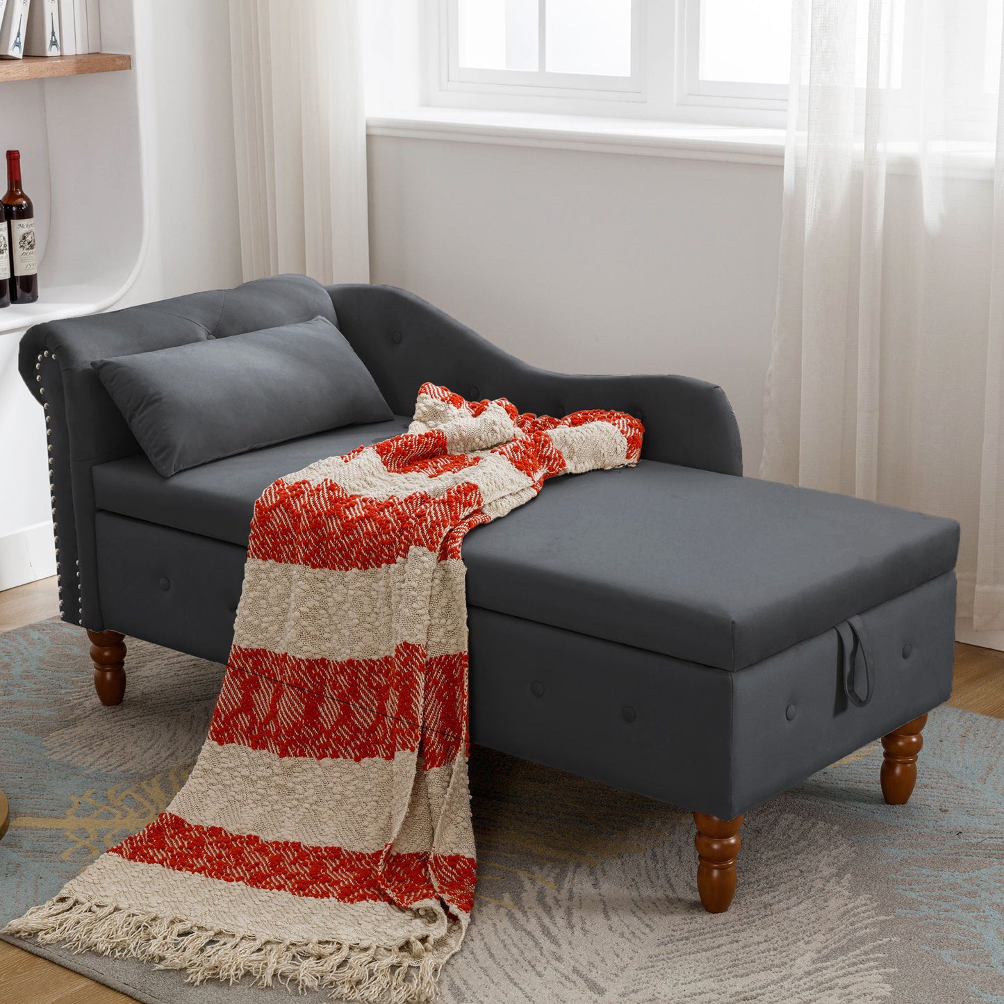 A modern dark gray Chaise Lounge Indoor, featuring velvet upholstery, nailhead trim, storage, and a pillow, is accented with a red and cream striped throw near a window with sheer curtains in a cozy room.