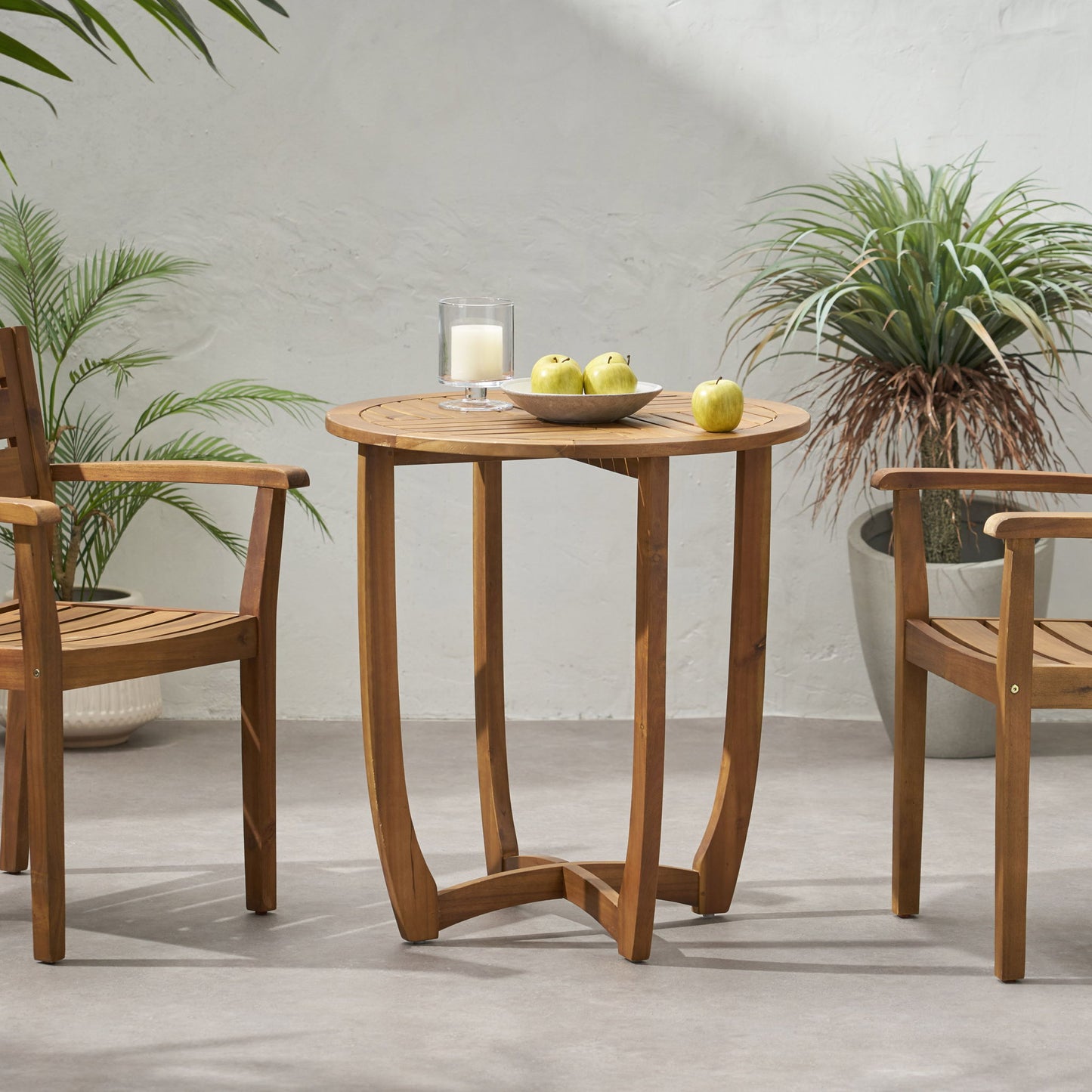 The Outdoor Circular Wood Bistro Table With Curved Legs And Slat Panel Table Top - Teak is placed between two wooden chairs, topped with a bowl of green apples and a candle, with potted plants on the patio against a light wall.
