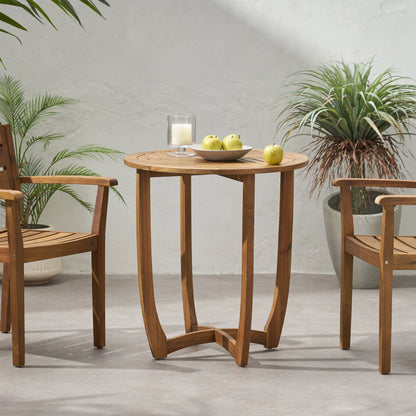 The Outdoor Circular Wood Bistro Table With Curved Legs And Slat Panel Table Top - Teak is placed between two wooden chairs, topped with a bowl of green apples and a candle, with potted plants on the patio against a light wall.