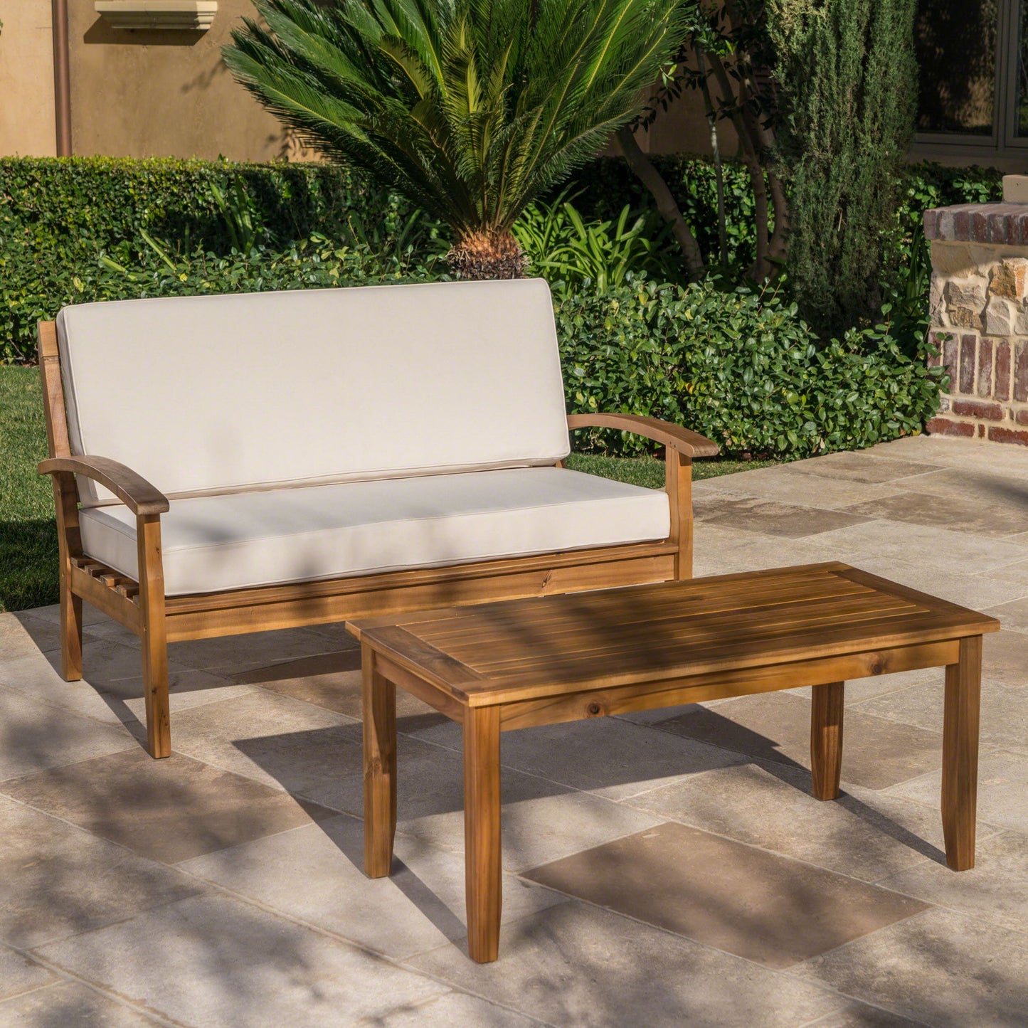 An Outdoor Chat Set With Coffee Table And Loveseat sits on a stone patio with green plants and shrubs, featuring light cushions and matching wooden pieces.