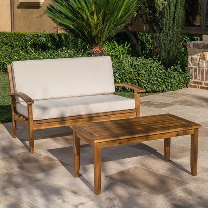 An Outdoor Chat Set With Coffee Table And Loveseat sits on a stone patio with green plants and shrubs, featuring light cushions and matching wooden pieces.