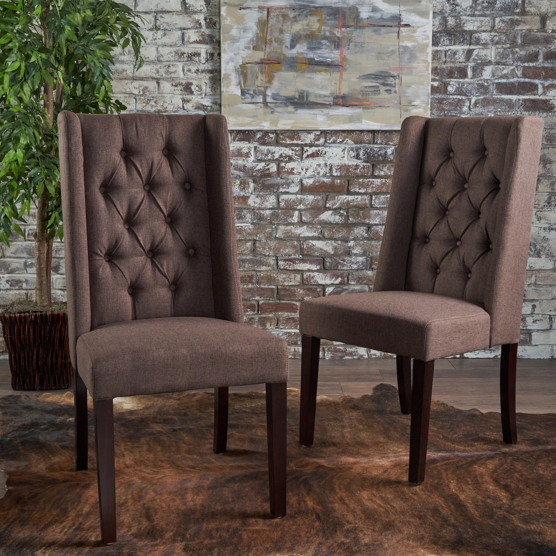 Two Sophisticated Tufted Dining Chairs with high back designs and dark wooden legs sit on a brown rug before a whitewashed brick wall, complemented by a potted plant and abstract art in the background.