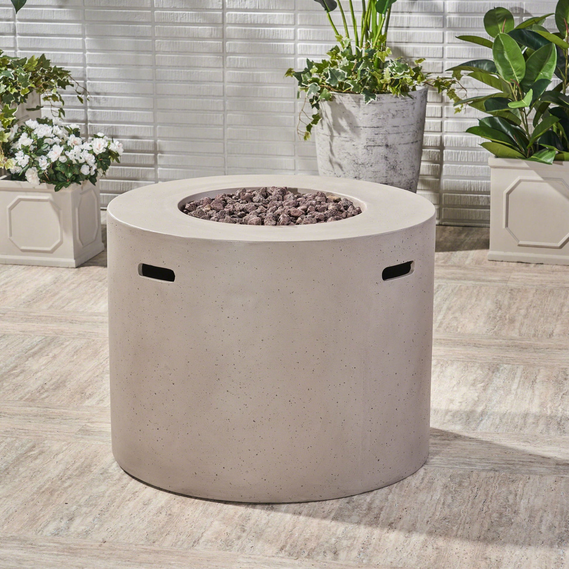 A Round Iron Fire Pit With 40,000 Btu sits on a patio with black stones on top, surrounded by green potted plants and a white tiled wall in the background.