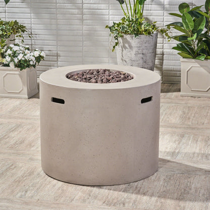 A Round Iron Fire Pit With 40,000 Btu sits on a patio with black stones on top, surrounded by green potted plants and a white tiled wall in the background.