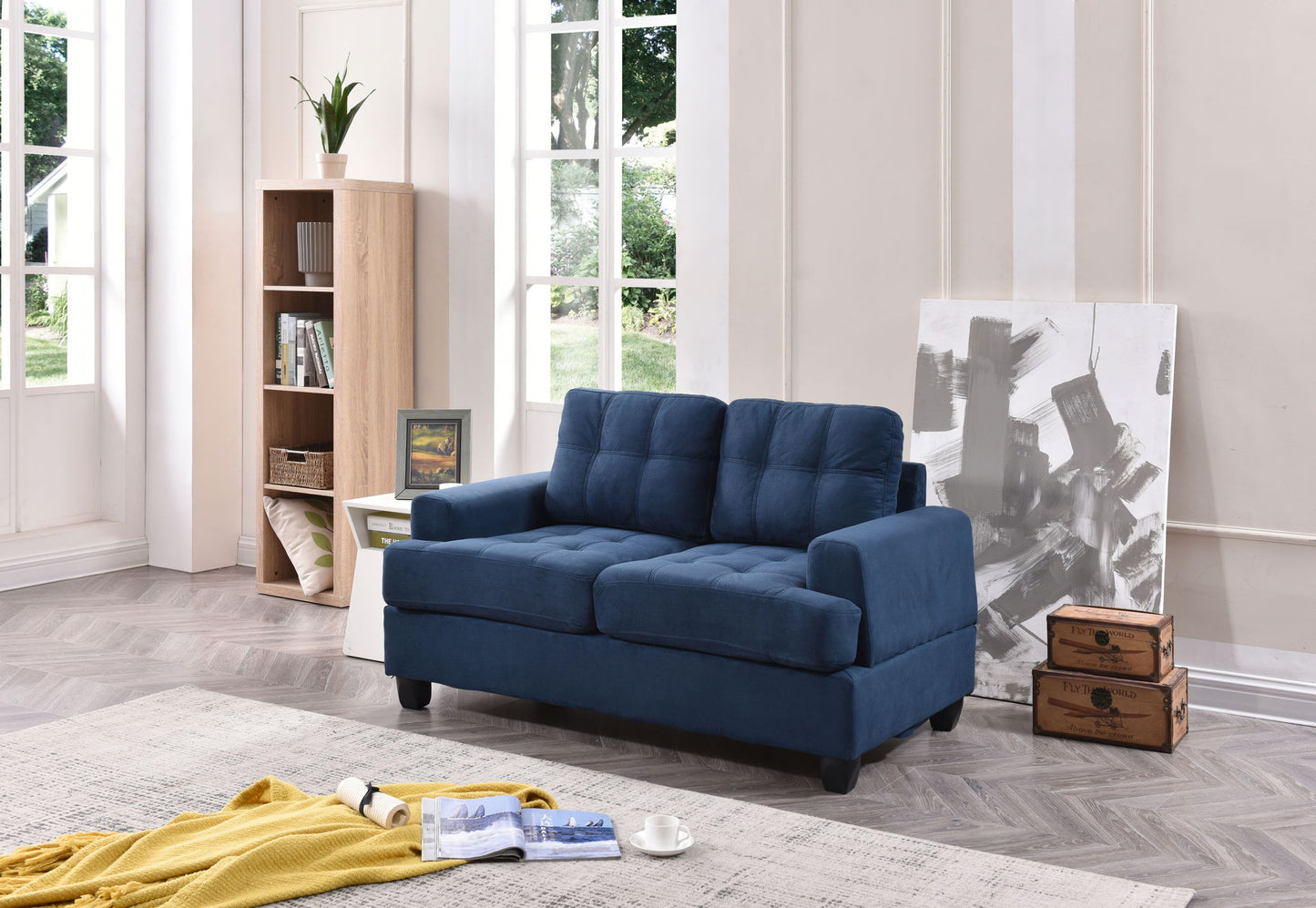 A modern living room featuring the Stylish Transitional Loveseat For Two in blue, a bookshelf, abstract painting, vintage suitcases, large sunlit windows, plus a cosy rug and throw blanket with a magazine on the floor.