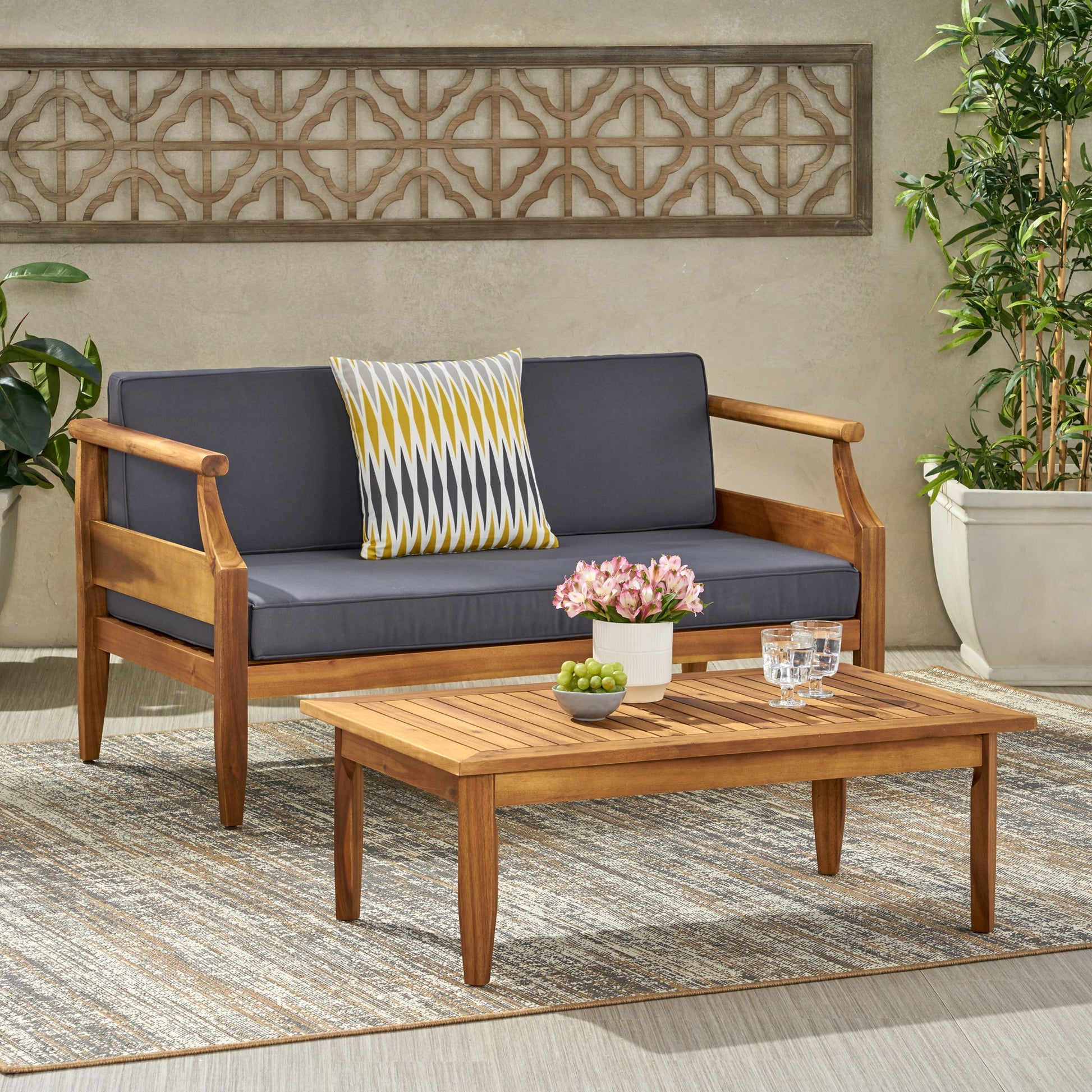 The Aston - 2 Piece Wood Outdoor Patio Conversation Seating Set with Cushions features a wooden sofa with dark gray cushions, a patterned pillow, and a matching coffee table adorned with flowers, grapes, two glasses of water, and surrounded by potted plants.