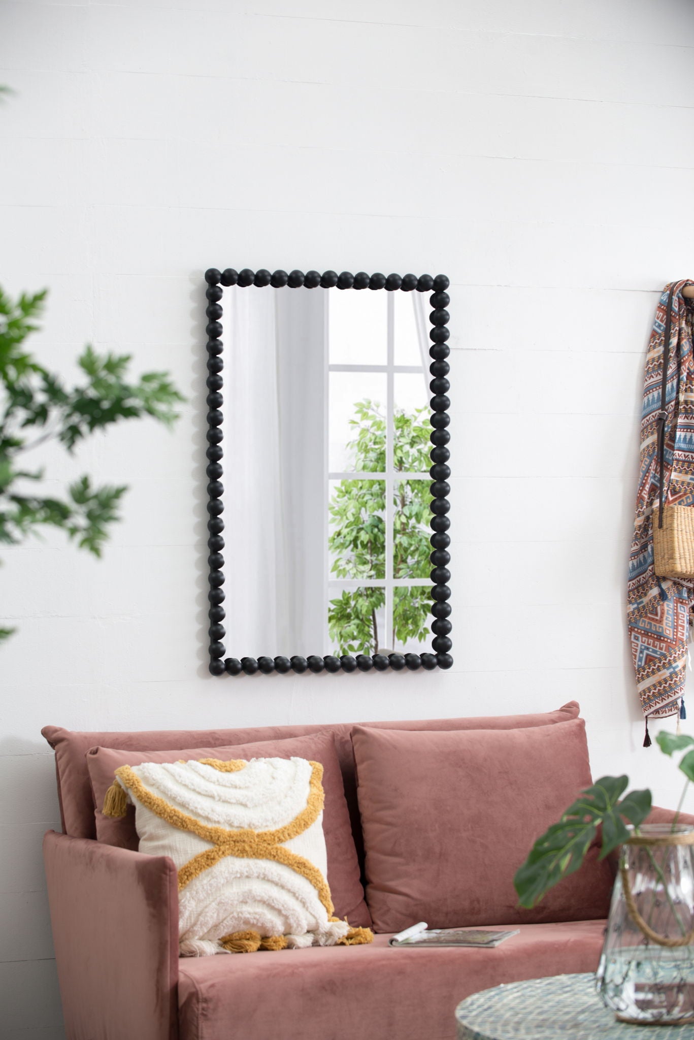 A Rectangle Mirror, Wall Mirror For Living Room Bedroom Entryway - Black hangs on a white wall above a pink velvet sofa with a patterned pillow. Nearby are a patterned scarf, wicker bag, and green plants that brighten the room.