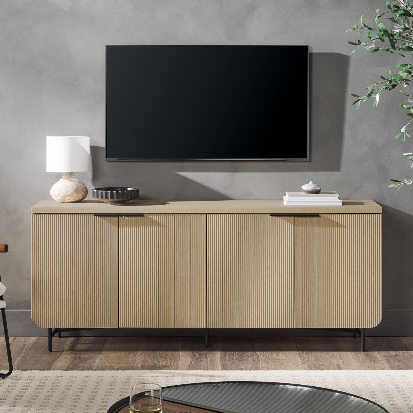 A flat-screen TV is mounted on a gray wall above the Modern Fluted Door Minimalist Storage Sideboard, which displays a white lamp, a round dark tray, and stacked books. A nearby plant and table are also visible.
