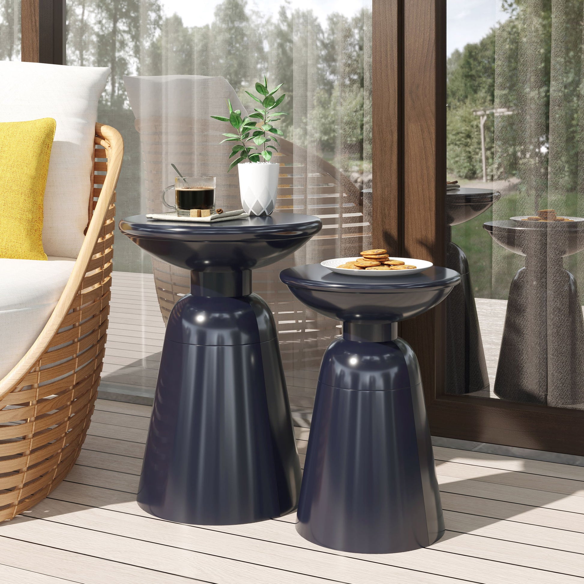Two Serenita Outdoor Side Tables with a flared design, shown in dark blue on a light wooden patio. One holds a plant and coffee, the other a plate of cookies. A wicker chair with yellow cushions and large windows complete the scene.