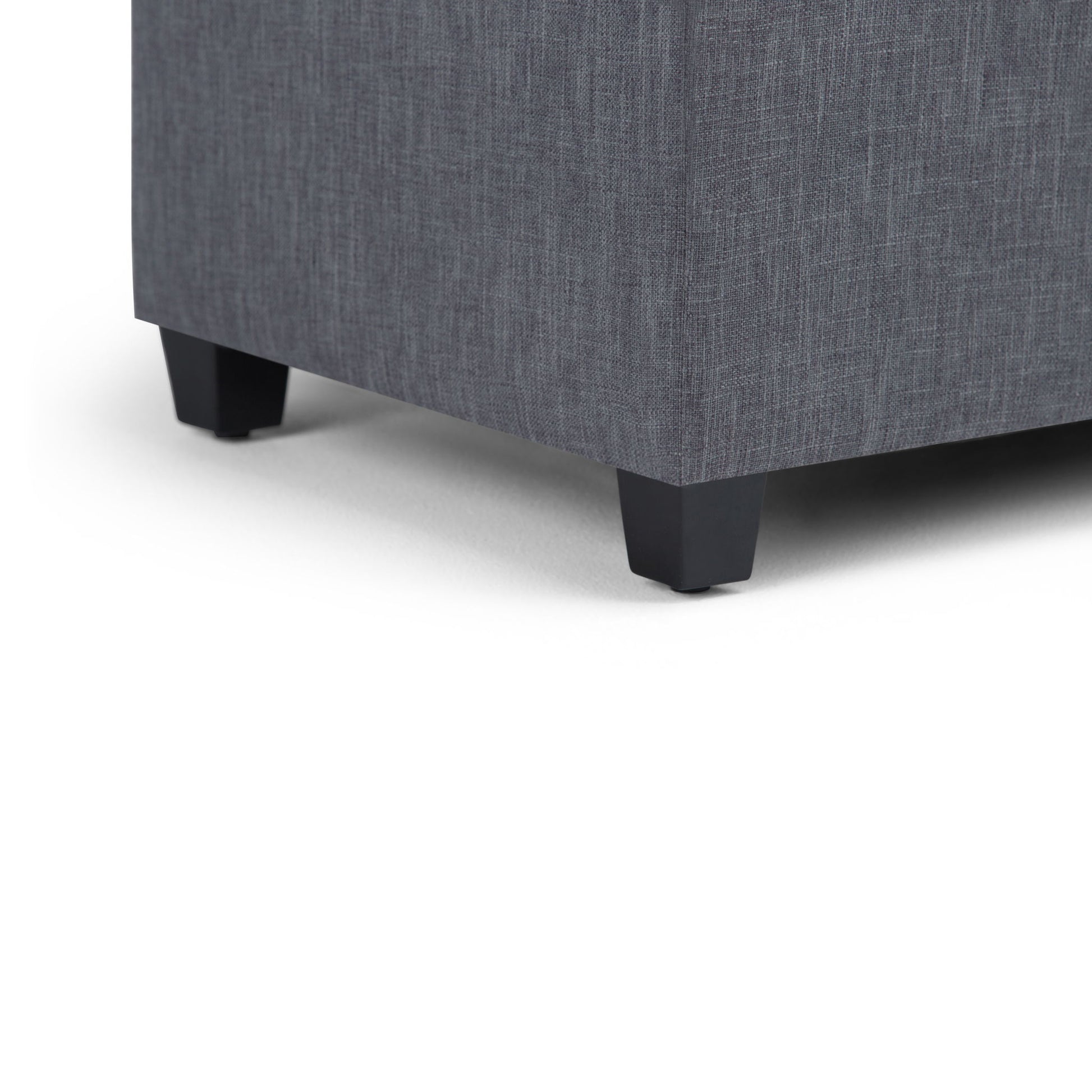 Close-up of the lower corner of the Sienna - Upholstered Storage Ottoman Bench, featuring gray textured fabric and short black legs, resting on a white surface.