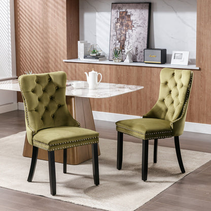 A set of two Nikki modern velvet-upholstered dining chairs with tufting and nailhead trim sit on a beige rug beside a marble table, framed by wood-paneled walls, shelves, and decorative accents in the dining area.