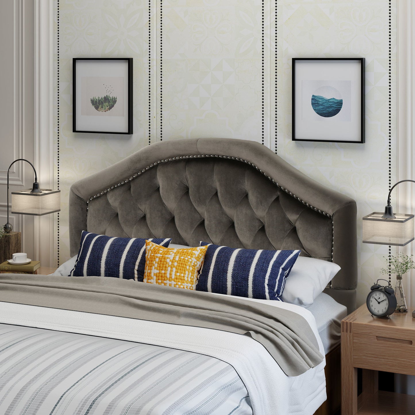 A neatly made bed features the Kaarina Contemporary Tufted Fabric Headboard in gray, with decorative pillows and striped bedding. Two framed pictures hang above, while nightstands with lamps and an alarm clock flank the bed.