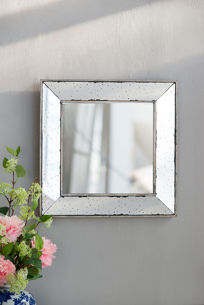 The Distressed Accent Mirror, a traditional style framed wall mirror for living room, hangs on a light gray wall. A vase with pink flowers and greenery appears in the lower left corner as sunlight casts soft shadows nearby.