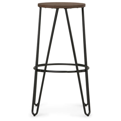 The Simeon Multifunctional Metal Stool features a tall, minimalist design with a round wood seat, black metal hairpin legs, and a circular footrest connecting the legs near the base.