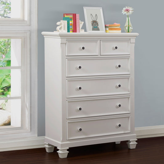The Glendale 6 Drawer Chest in white stands against a gray wall by a window. Its top displays books, letter decor, a framed bunny print, a vase with flowers, and small items.