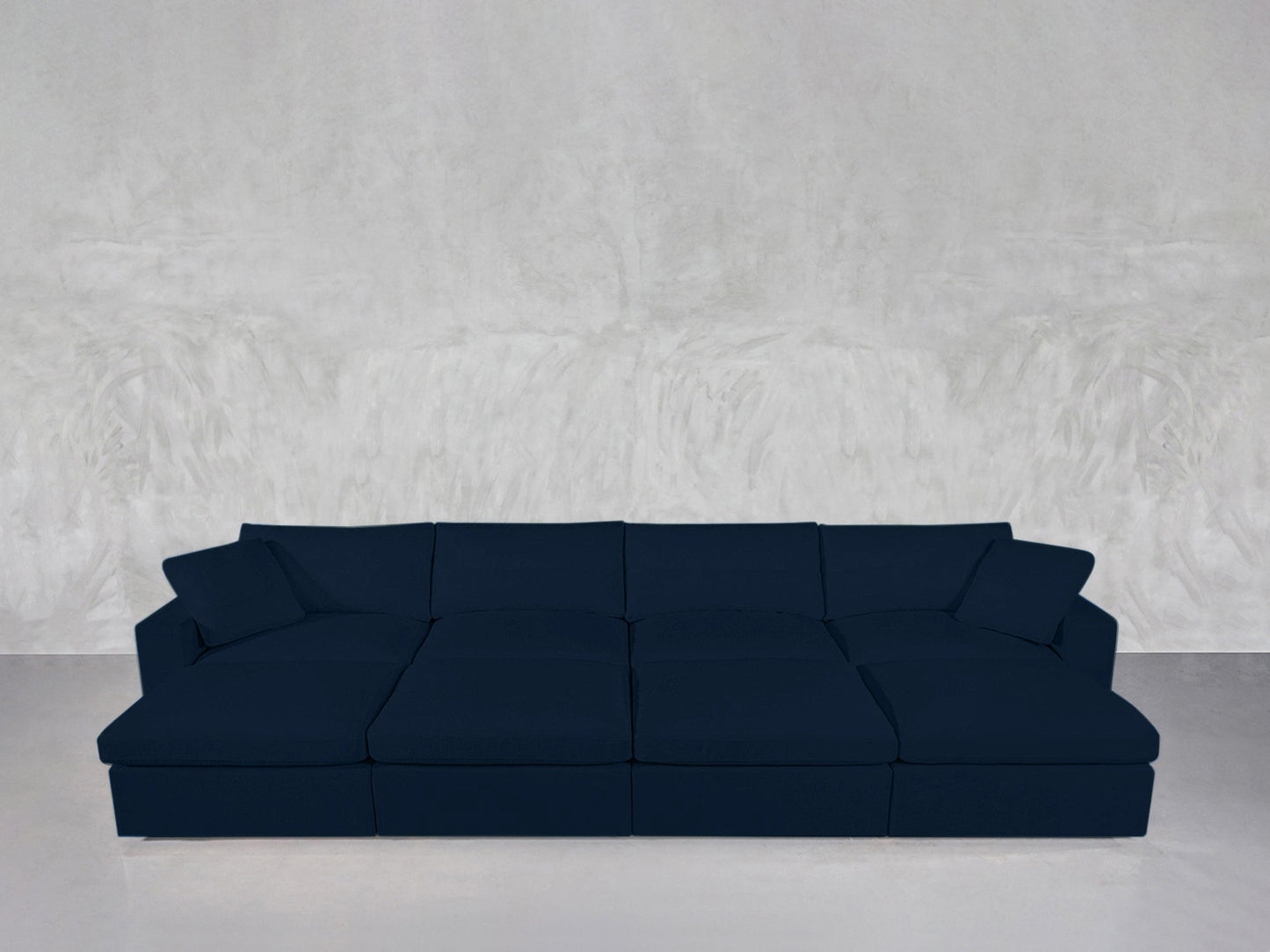 An 8-Seat Modular Daybed in dark blue with plush cushions and pillows sits against a light gray textured wall on a smooth, light-colored floor.