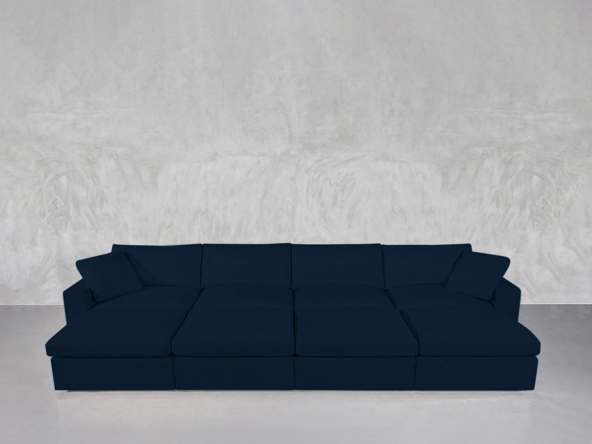 An 8-Seat Modular Daybed in dark blue with plush cushions and pillows sits against a light gray textured wall on a smooth, light-colored floor.