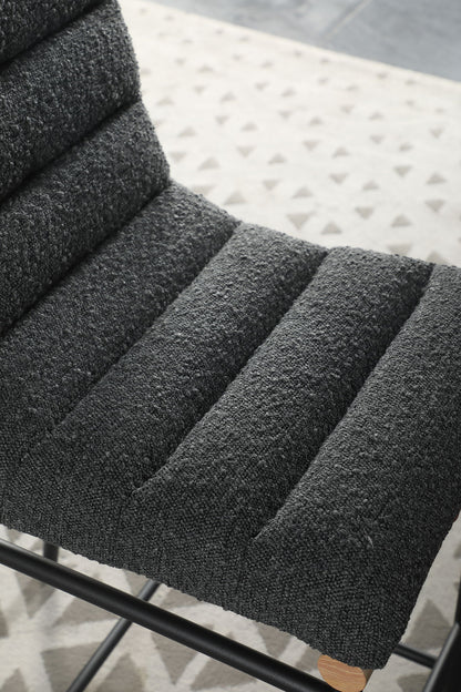 Close-up of the Lahni 17" Boucle Fabric Counter Height Chair, featuring textured dark gray boucle upholstery, horizontal tufted padding, and a black metal frame. A patterned rug is seen in the background.
