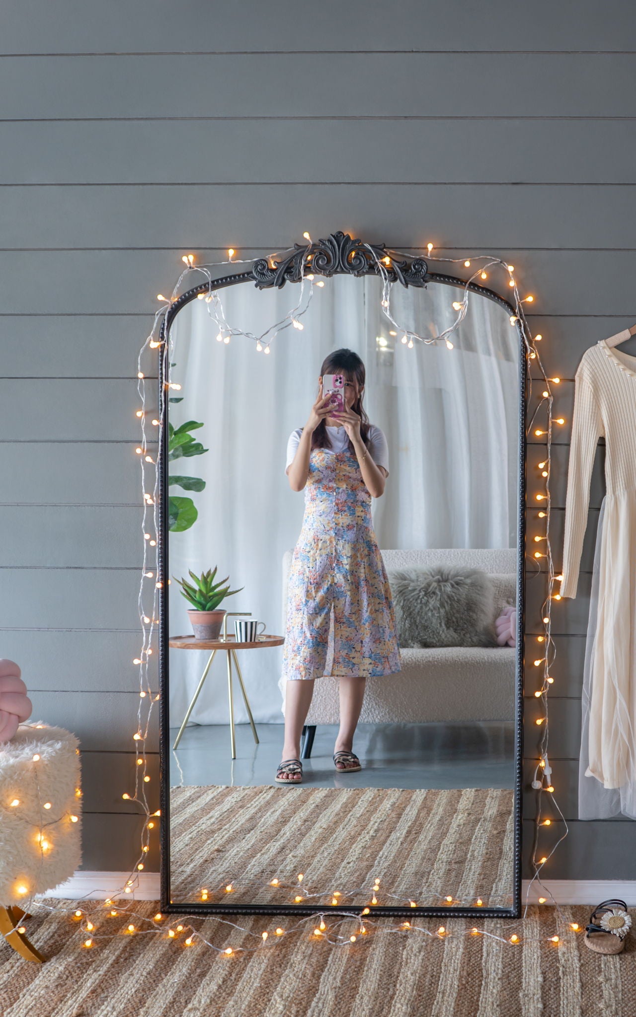 A woman in a floral dress takes a mirror selfie in a cozy room, featuring the Arch Mirror, Baroque Inspired Wall Decor—adorned with string lights—with a plant, furry chair, and beige dress visible in the background.
