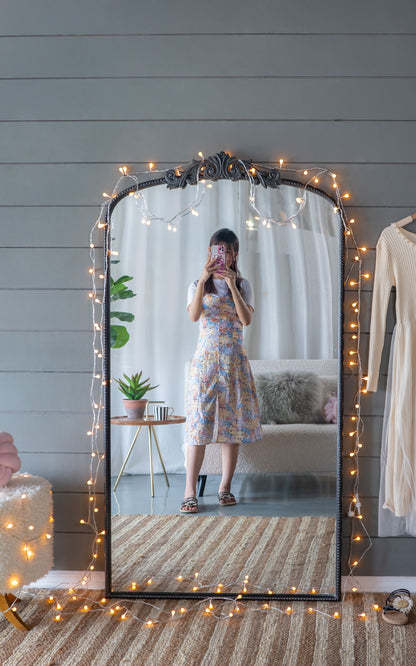 A woman in a floral dress takes a mirror selfie in a cozy room, featuring the Arch Mirror, Baroque Inspired Wall Decor—adorned with string lights—with a plant, furry chair, and beige dress visible in the background.