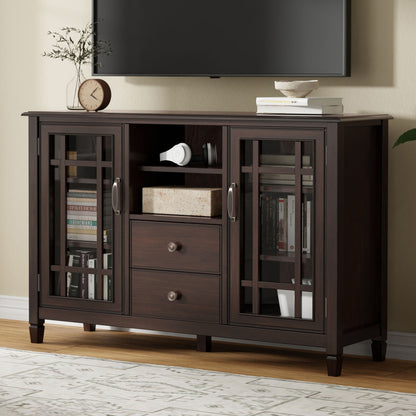 The Connaught Tall TV Stand in Brown features glass-paneled doors, two center drawers, and an open shelf. Books, decor, and electronics are neatly arranged inside and on top. A TV is wall-mounted above it against a beige background.