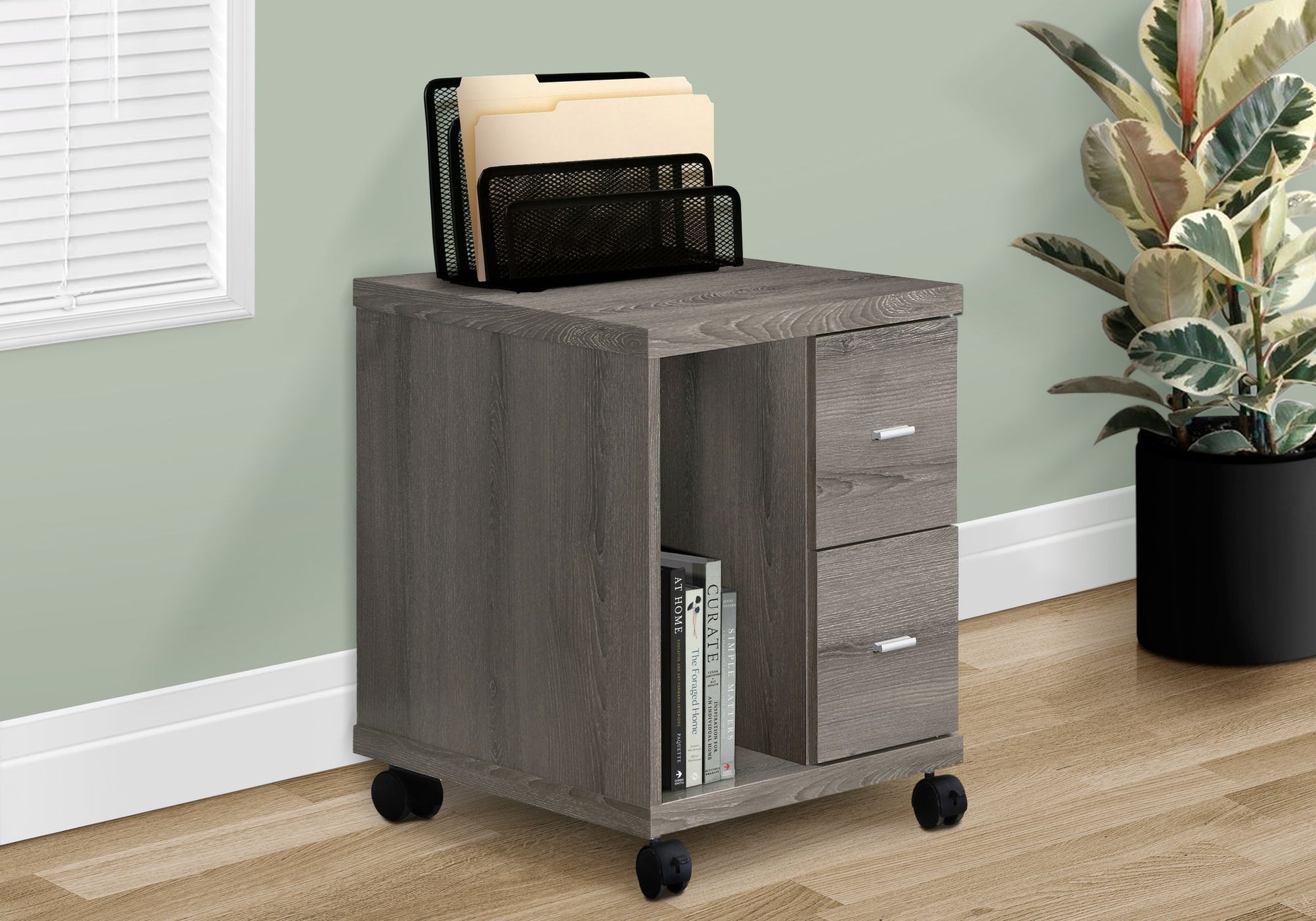 A contemporary gray wooden rolling file cabinet with two drawers and an open shelf for books sits by a plant and window on a wood floor. A black mesh file organizer with folders rests on top of this modern mobile storage unit.