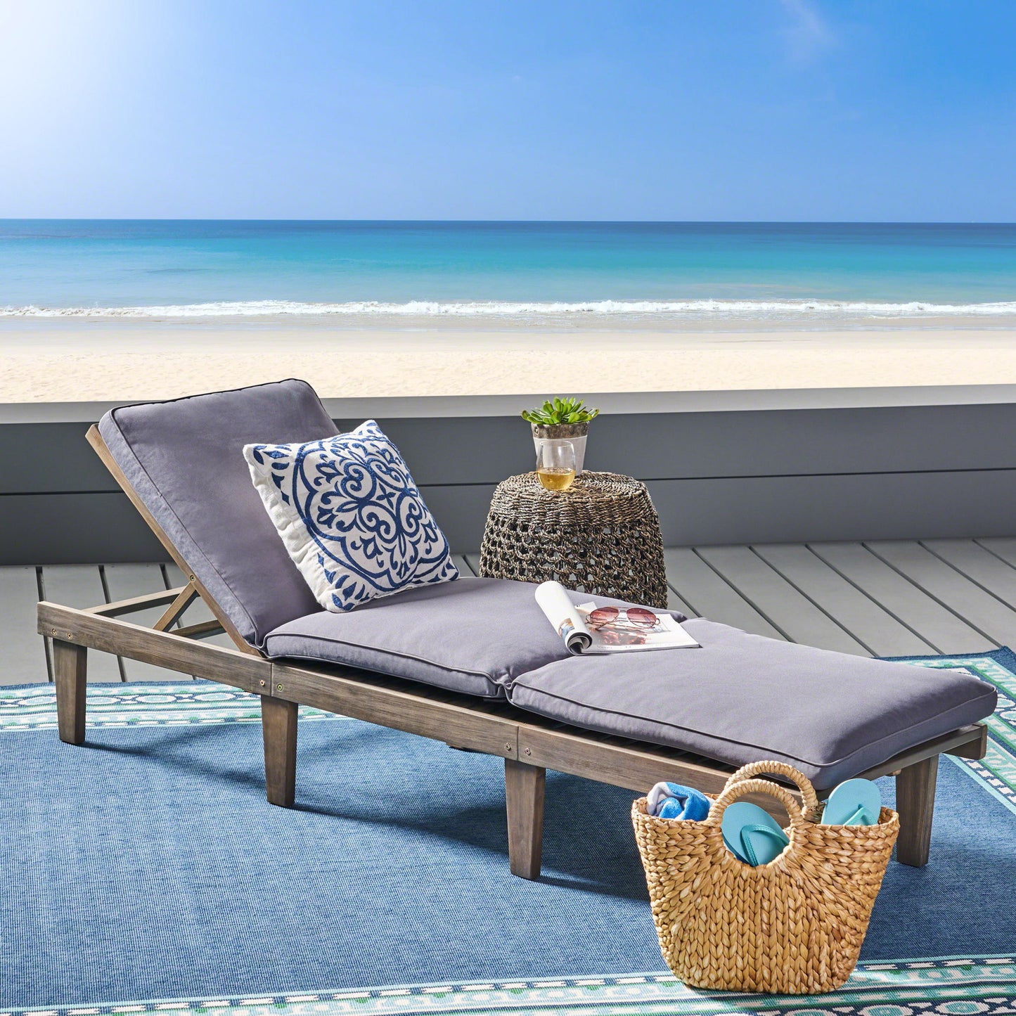The Ariana - Wood Chaise Lounge With Cushion, featuring gray cushions and a decorative pillow, sits on a blue outdoor rug near the beach. A small round table holds a drink and magazine, with a straw tote bag nearby and the ocean in view.