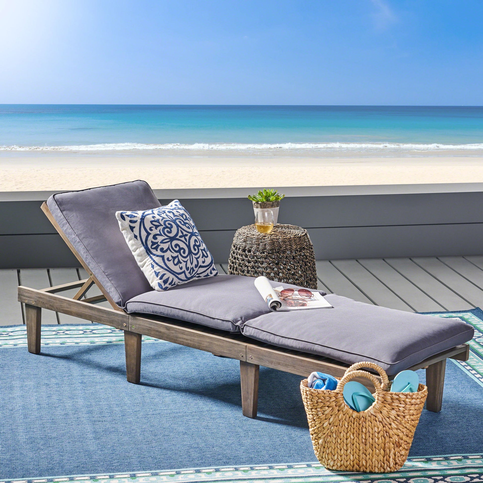 The Ariana - Wood Chaise Lounge With Cushion, featuring gray cushions and a decorative pillow, sits on a blue outdoor rug near the beach. A small round table holds a drink and magazine, with a straw tote bag nearby and the ocean in view.