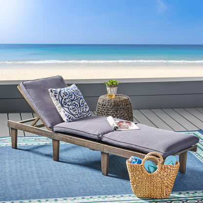 The Ariana - Wood Chaise Lounge With Cushion, featuring gray cushions and a decorative pillow, sits on a blue outdoor rug near the beach. A small round table holds a drink and magazine, with a straw tote bag nearby and the ocean in view.