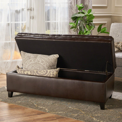 The Juliana Storage Ottoman With Birch Legs, shown in dark brown leather with its lid open to reveal two patterned pillows, sits in a bright room with large windows, a plant, and part of a sofa nearby.