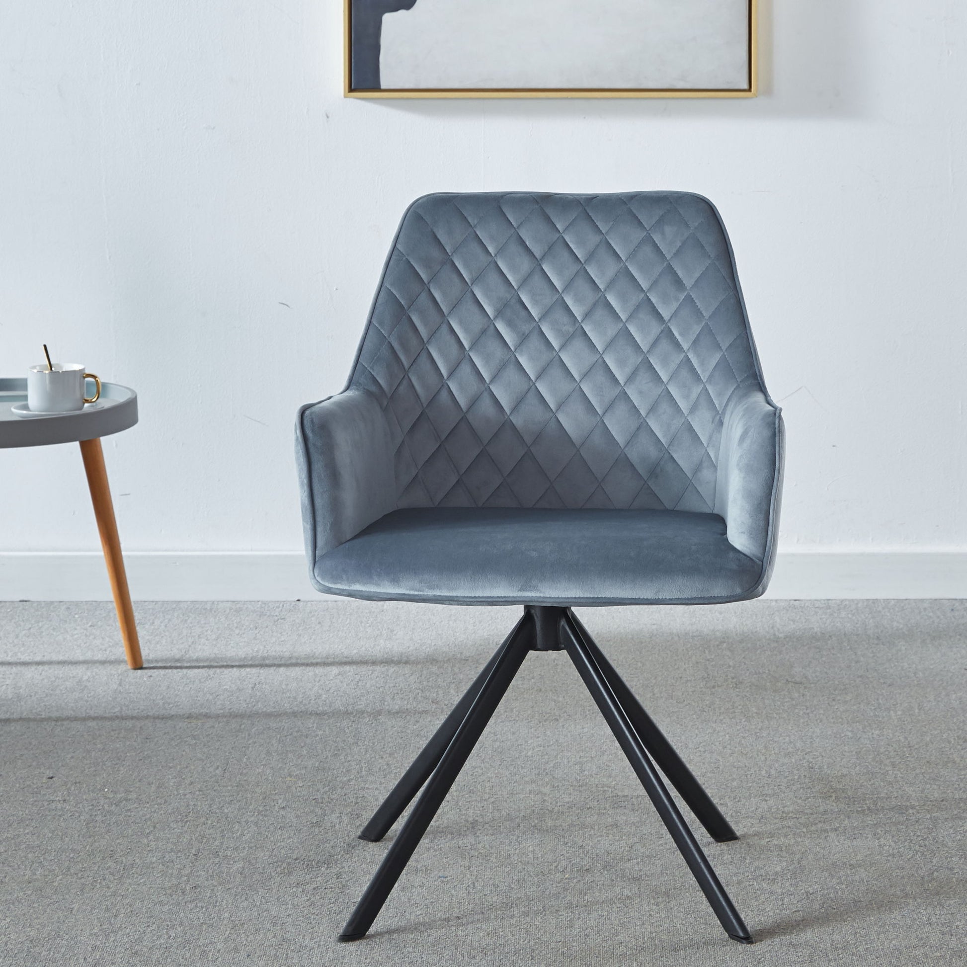 A Modern Desk Chair No Wheel, upholstered in blue with ergonomic design and metal legs, sits on a light gray carpet. To its left is a small round table with a coffee cup, and framed artwork hangs on the white wall behind.