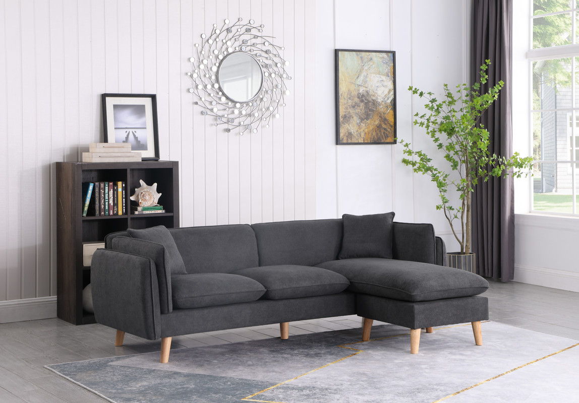 A modern living room featuring the Brayden Fabric Sectional Sofa Chaise in dark gray, a gray and white rug, a black bookshelf with books and decor, wall art, a round mirror, a green potted plant, and large windows with sheer curtains.