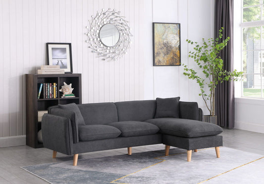 A modern living room featuring the Brayden Fabric Sectional Sofa Chaise in dark gray, a gray and white rug, a black bookshelf with books and decor, wall art, a round mirror, a green potted plant, and large windows with sheer curtains.