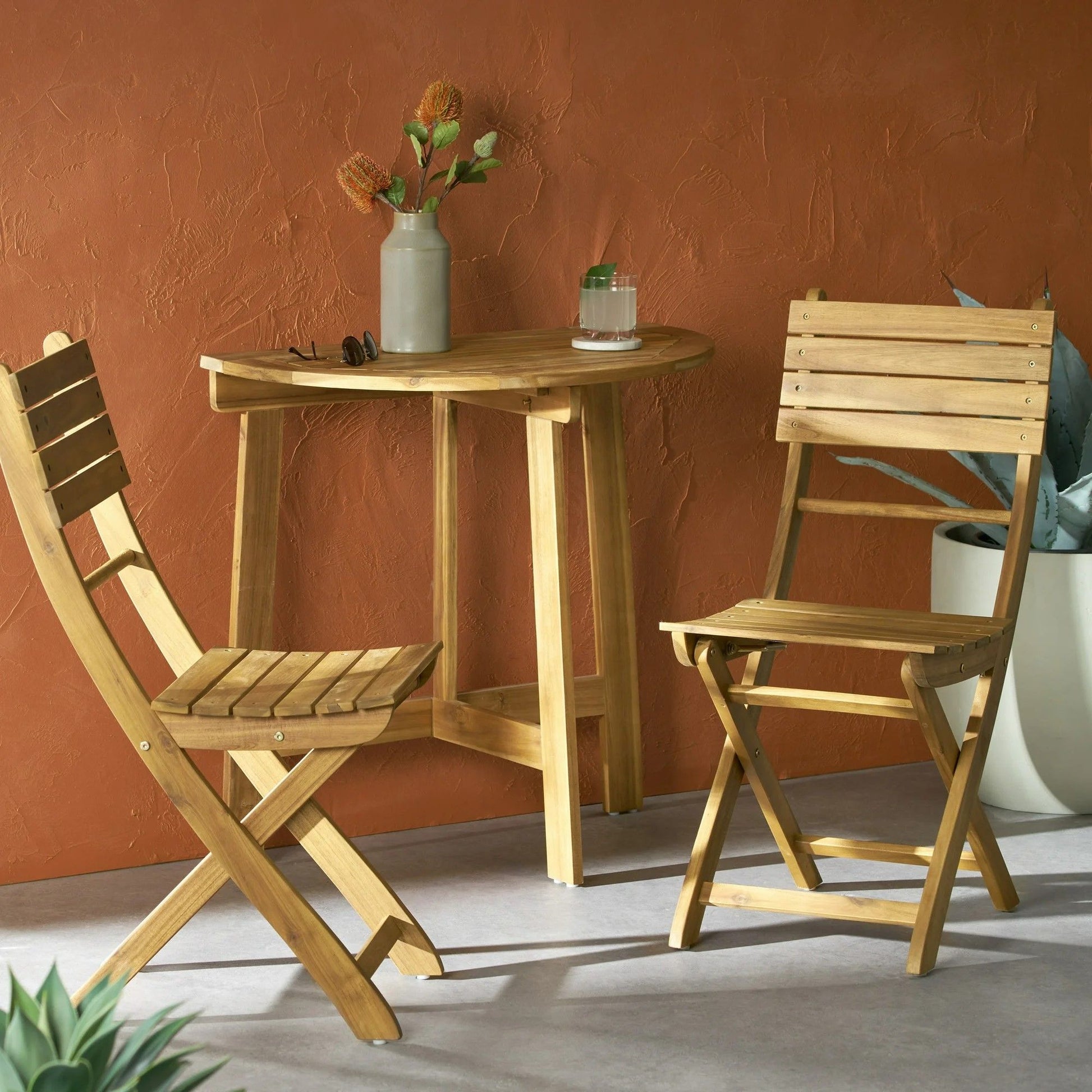 The Outdoor 3 Piece Half Round Folding Bistro Set, featuring two folding chairs and a table, sits on a gray floor against a terracotta wall. A vase with flowers and a glass rest on the table, with a large potted plant nearby.