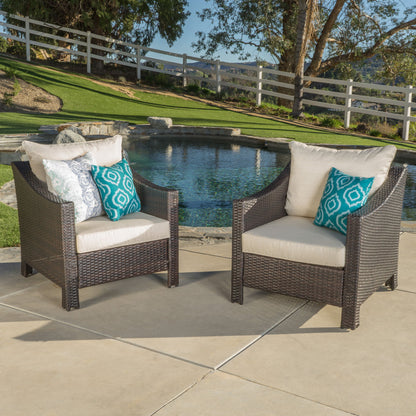 Two Antibes Armchairs with water resistant cream cushions and blue-patterned pillows sit on a concrete patio by a pool, overlooking a grassy lawn, trees, and a white fence.