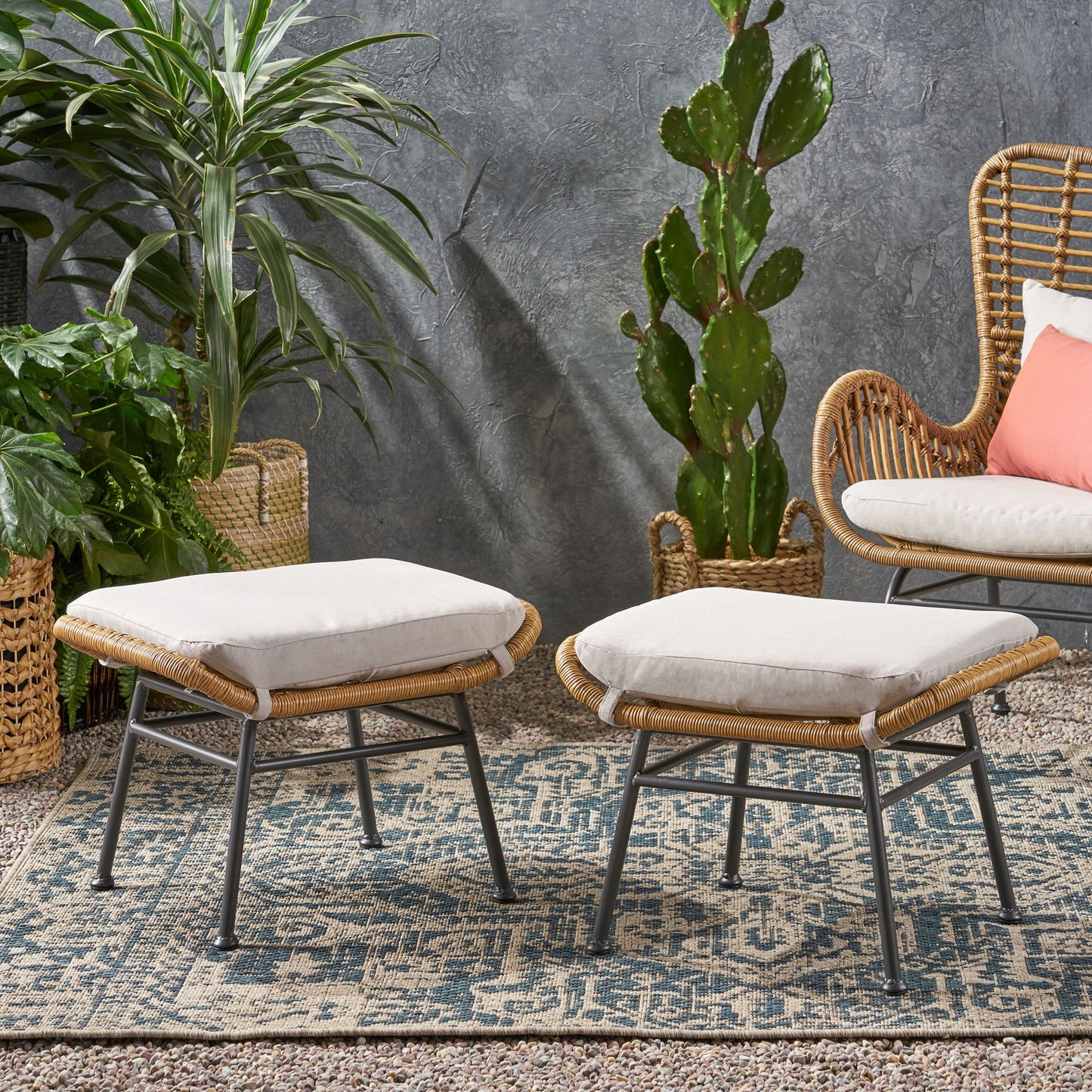 Two Outdoor Modern Boho Wicker Ottomans With Cushions (Set of 2) rest on a patterned rug next to a wicker chair with a coral pillow, surrounded by green potted plants and set against a textured gray wall.