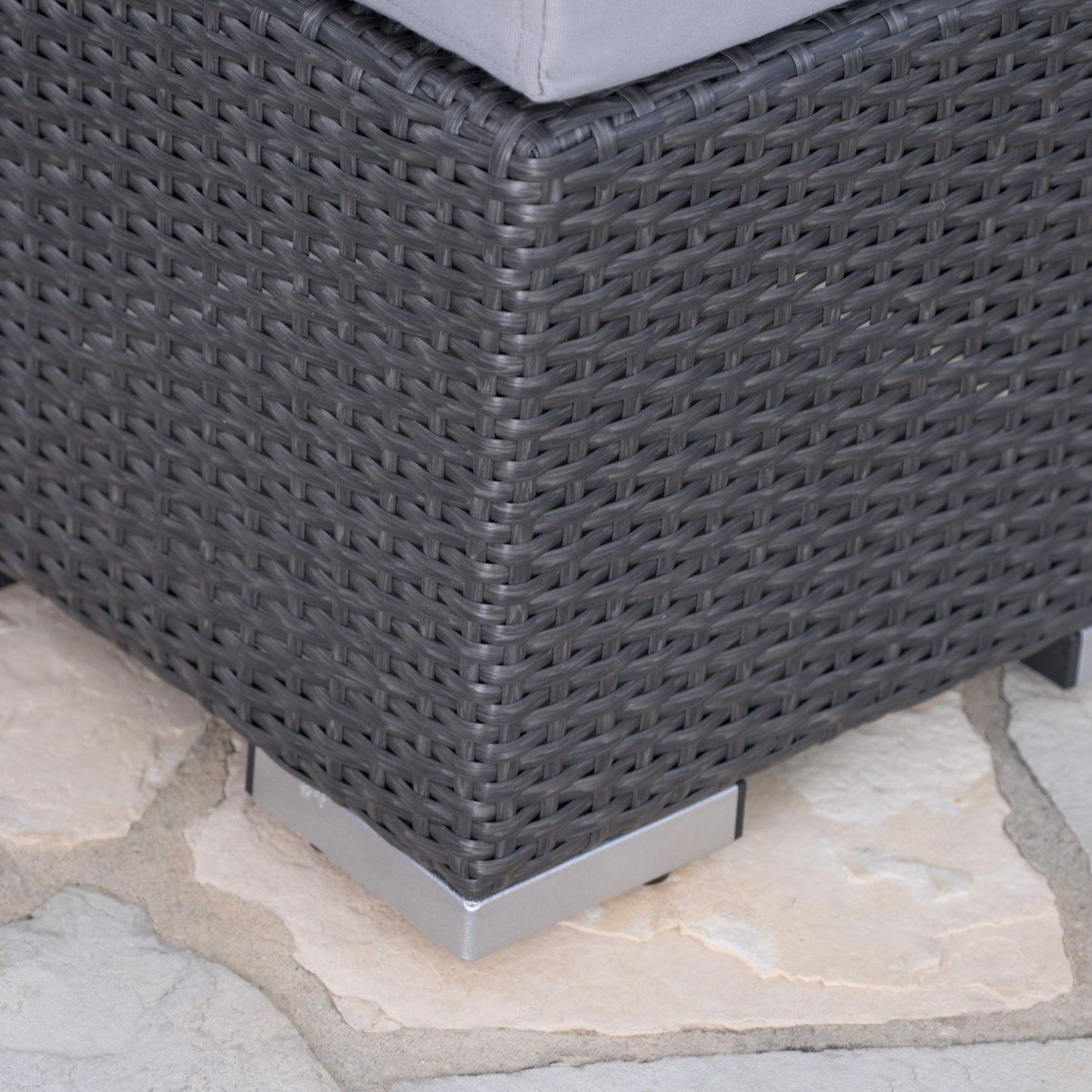 Close-up of the corner of the Santa Rosa - Outdoor Lounge Ottoman in dark gray wicker with a gray cushion, metal feet at the corners, and placed on a stone patio.