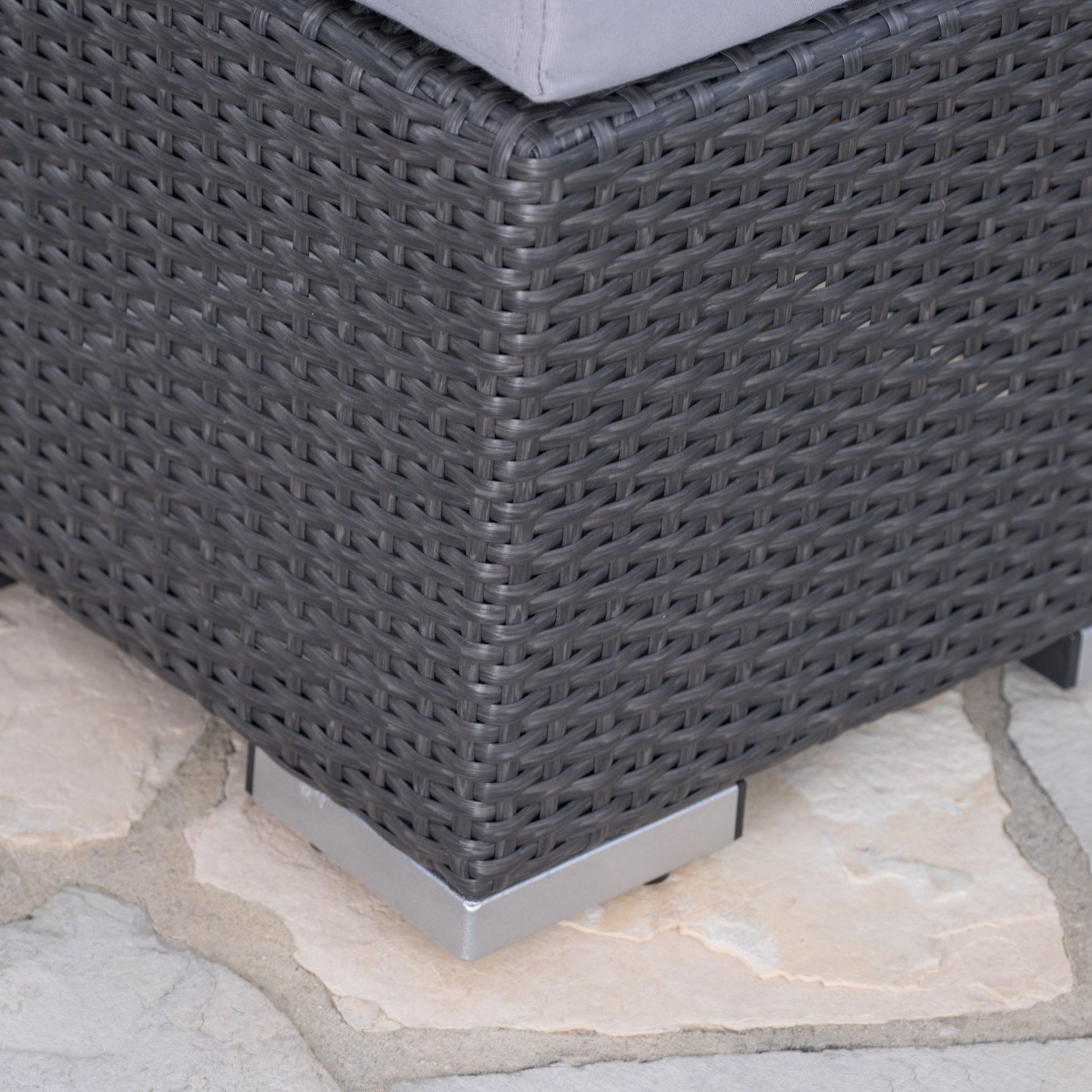 Close-up of the corner of the Santa Rosa - Outdoor Lounge Ottoman in dark gray wicker with a gray cushion, metal feet at the corners, and placed on a stone patio.