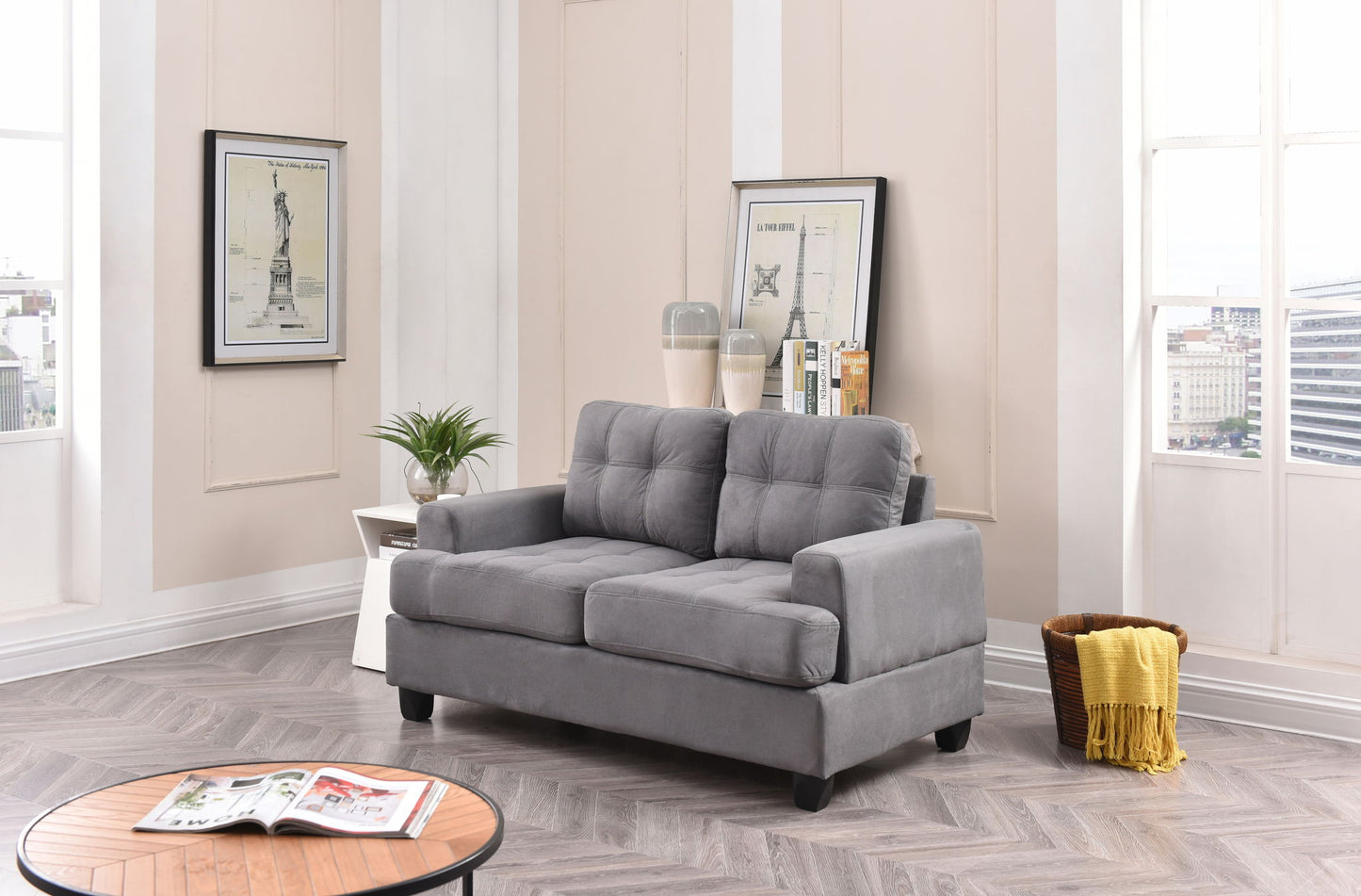 A modern living room featuring the Stylish Transitional Loveseat For Two in gray, light wood herringbone floors, a coffee table with magazines, white side table with a plant, framed art, books, and large windows with city views and yellow curtains.