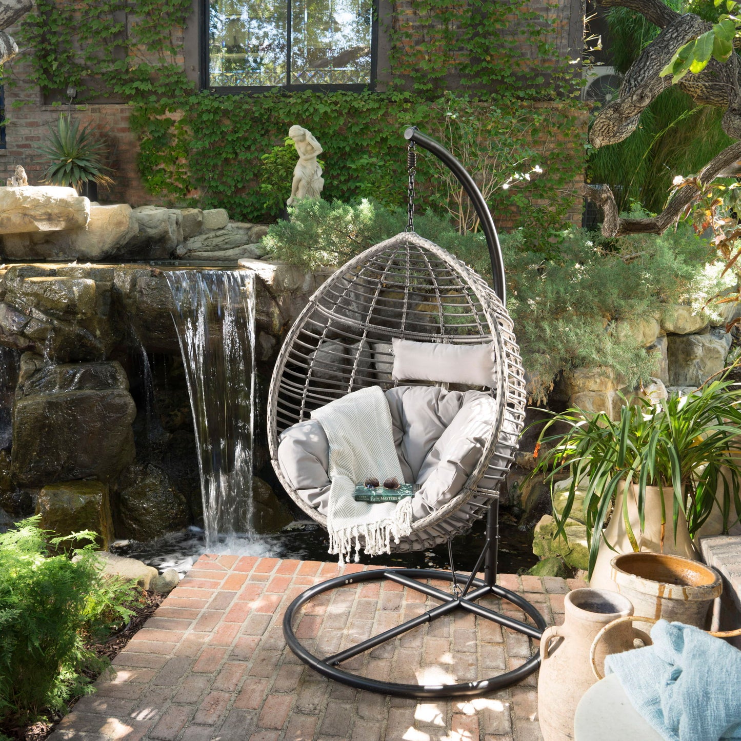 The Hanging Basket Chair With Metal Frame And Seat Cushion sits on a brick patio near a small waterfall, surrounded by lush greenery in a peaceful garden setting.