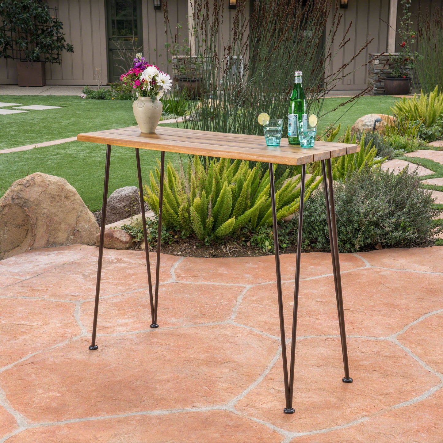 An Outdoor Bar Table With Wood Tabletop And Iron Hairpin Legs - Black / Teak stands on a stone patio, topped with a vase of flowers, a green bottle, and glasses with lime. Plants and garden landscaping appear in the background.