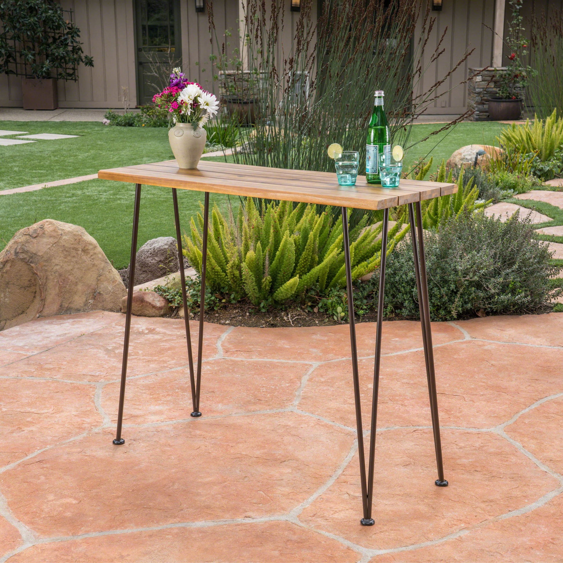 An Outdoor Bar Table With Wood Tabletop And Iron Hairpin Legs - Black / Teak stands on a stone patio, topped with a vase of flowers, a green bottle, and glasses with lime. Plants and garden landscaping appear in the background.