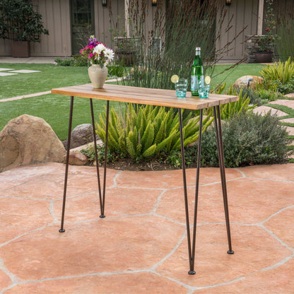 An Outdoor Bar Table With Wood Tabletop And Iron Hairpin Legs - Black / Teak stands on a stone patio, topped with a vase of flowers, a green bottle, and glasses with lime. Plants and garden landscaping appear in the background.
