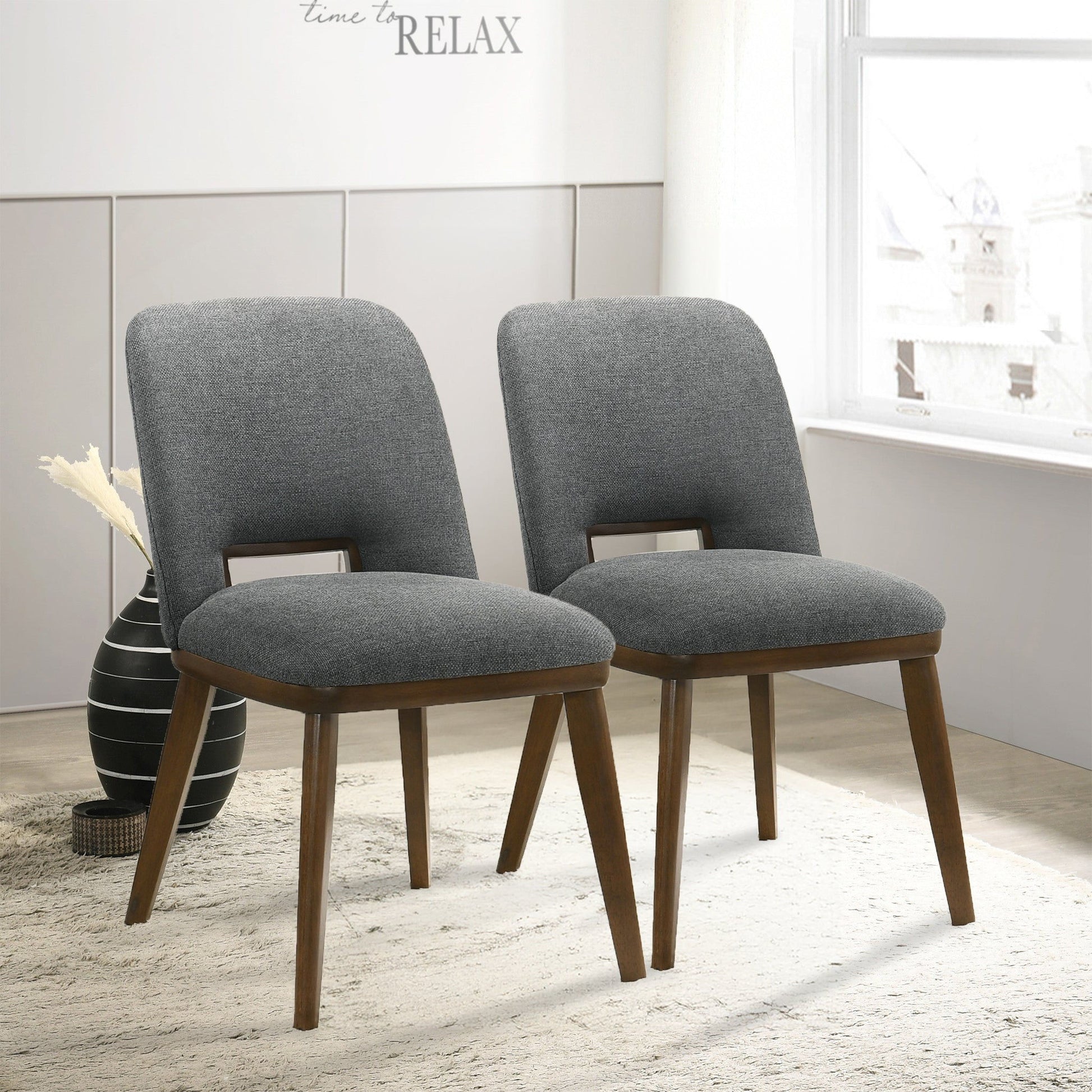 Two Blake Modern Dining Chairs (set of 2) with gray upholstery and wooden legs are on a light rug in a bright room near a window. A black vase with dried flowers sits in the background. "time to RELAX" is written on the wall.