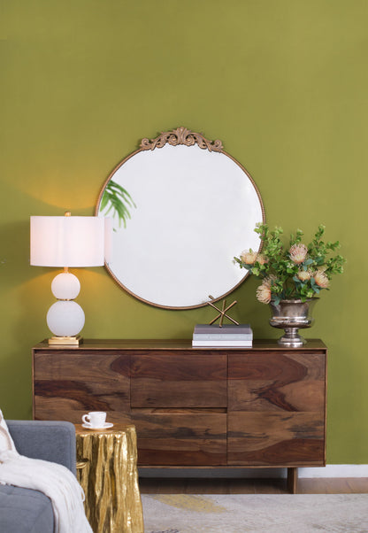A stylish wooden sideboard with a Classic Design Mirror with a Baroque-inspired frame, a white lamp, silver vase of green flowers, books, and a gold accent sits against an olive green wall. A gold side table and gray sofa are partly visible.