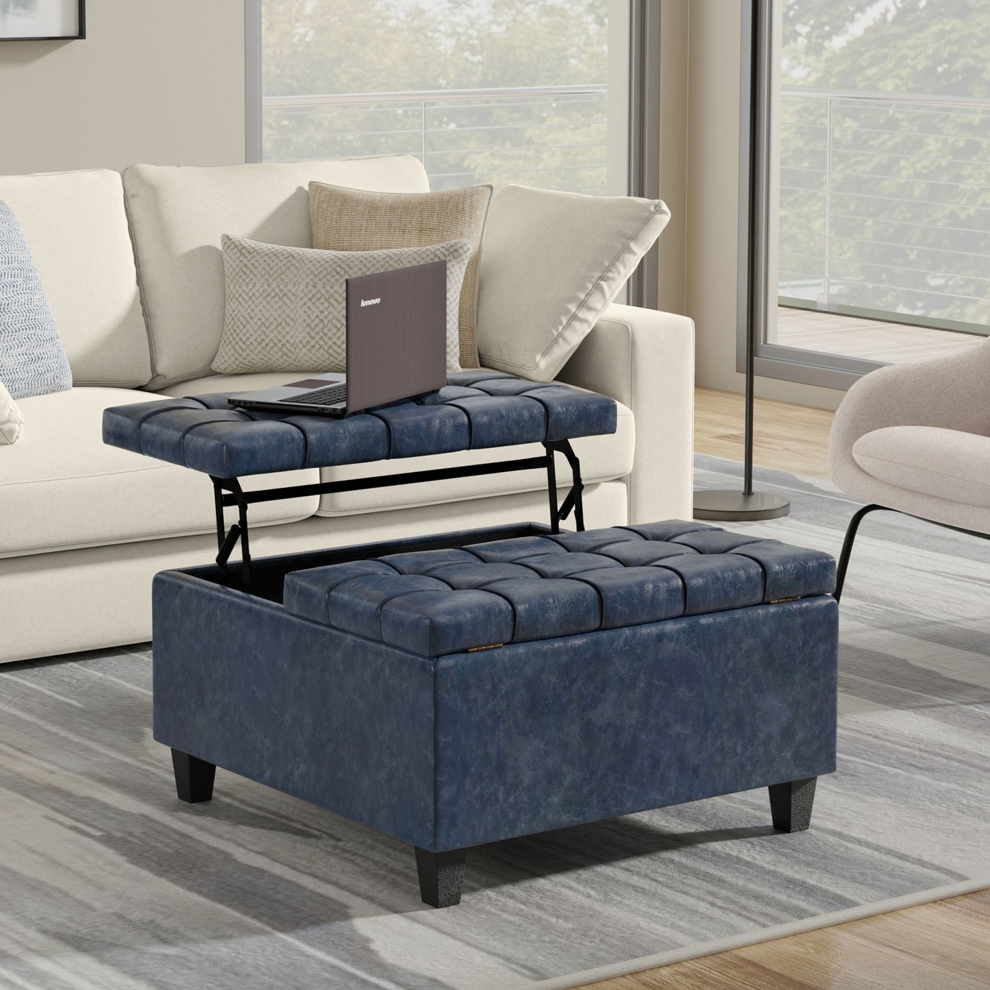 The Harrison Upholstered Coffee Table Storage Ottoman with a lift-top holds an open laptop in a modern living room featuring a cream sofa, cushions, and large windows.